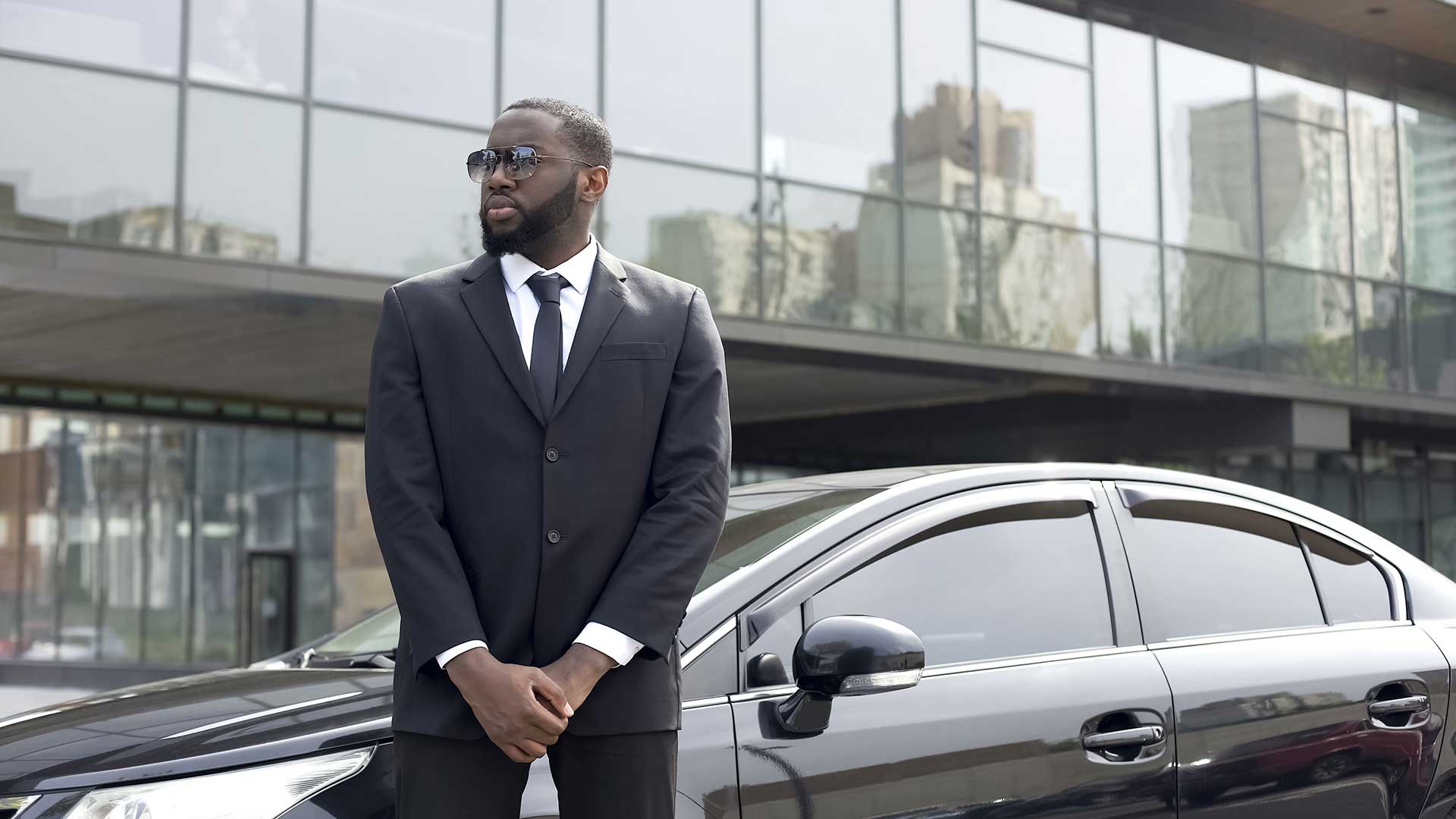 Bodyguard in a suit standing in front of a town car parked in front of an office building